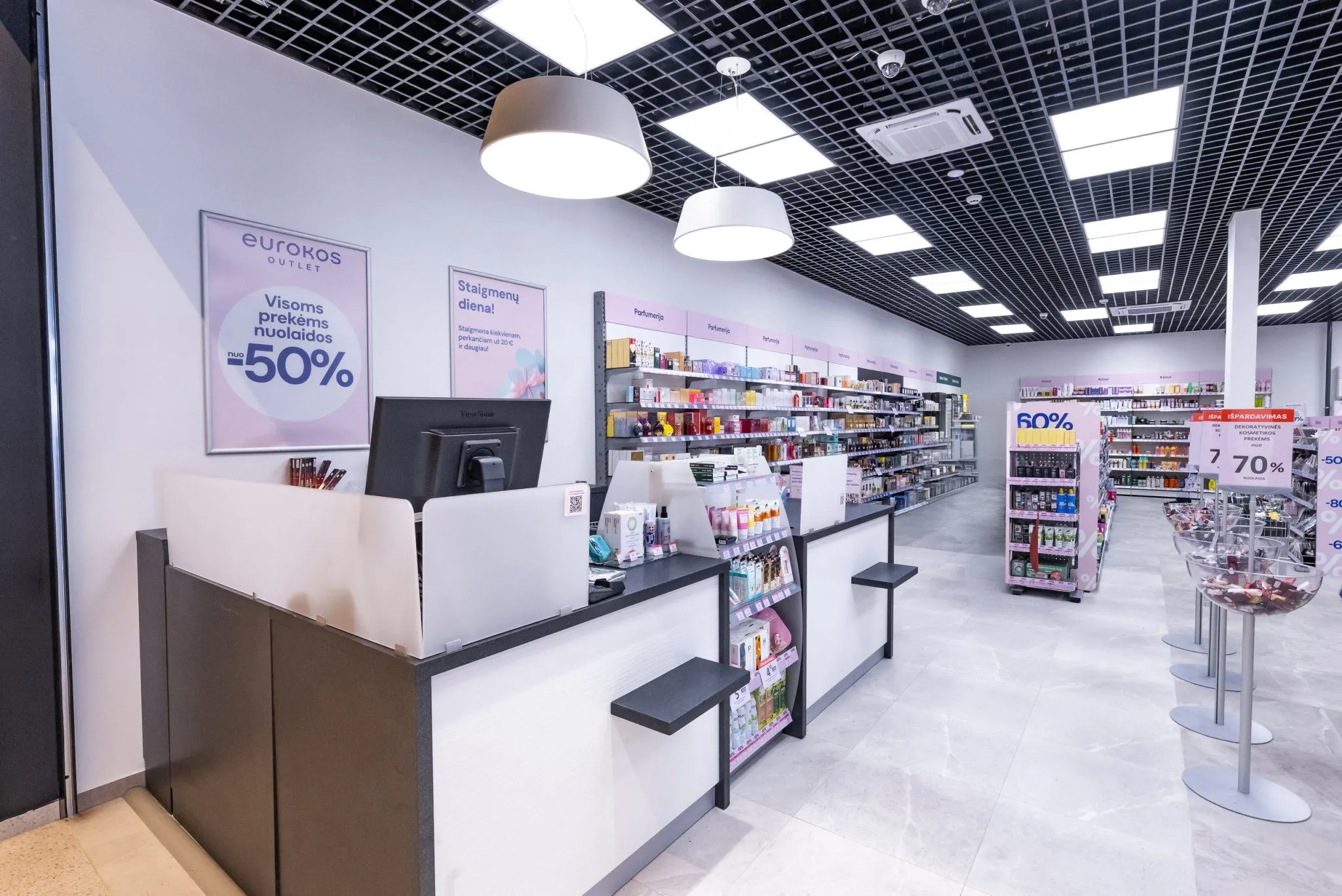 Eurokos Outlet parduotuvės vidus: kasa, plakatas „Visoms prekėms nuolaidos nuo -50 %“ ir lentynos su kvepalais
